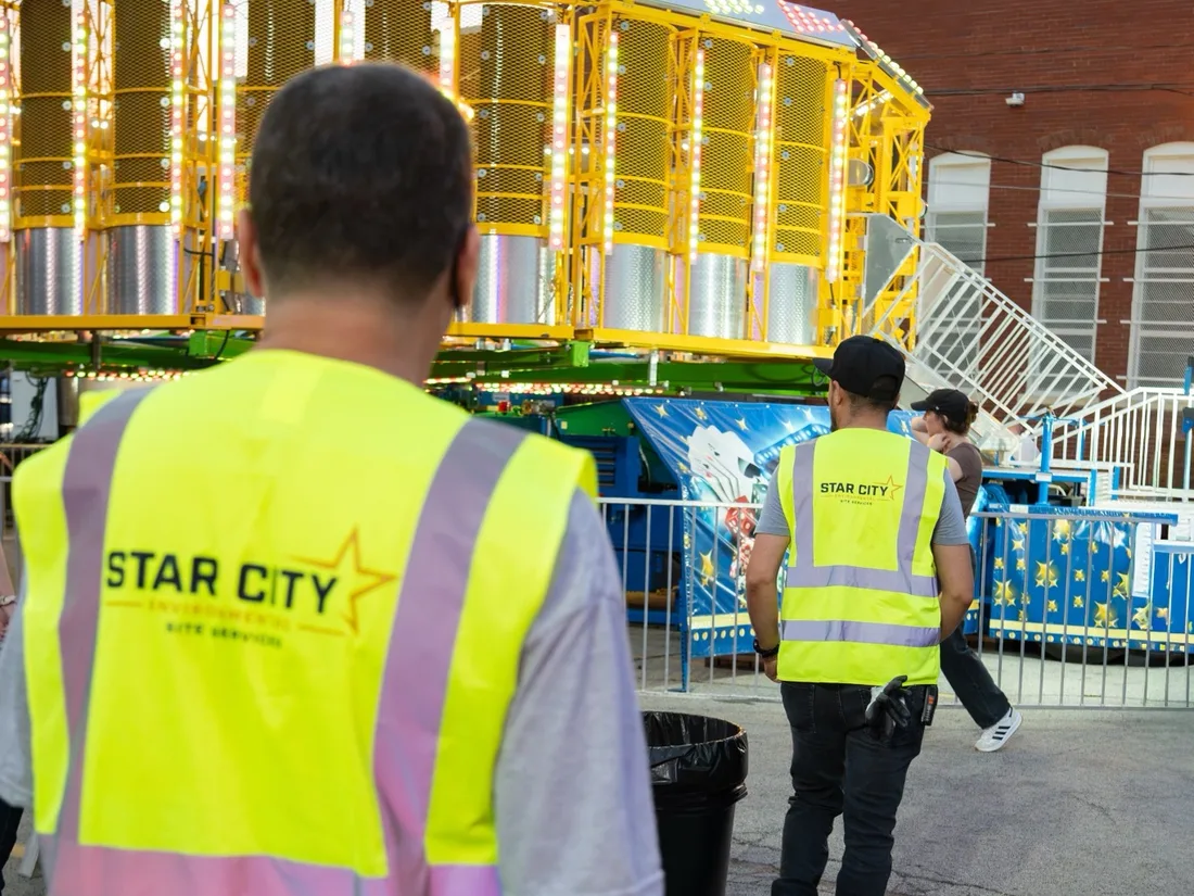 Two crew members in Star City Environmental vests working a midway at dusk, one walking a trash can past a lit ride.