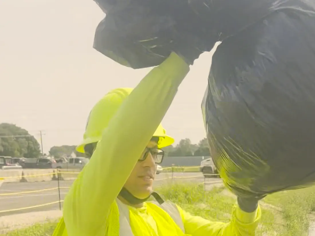 A Star City Environmental crew member in a hard hat lifting filled refuse bags into a dumpster.