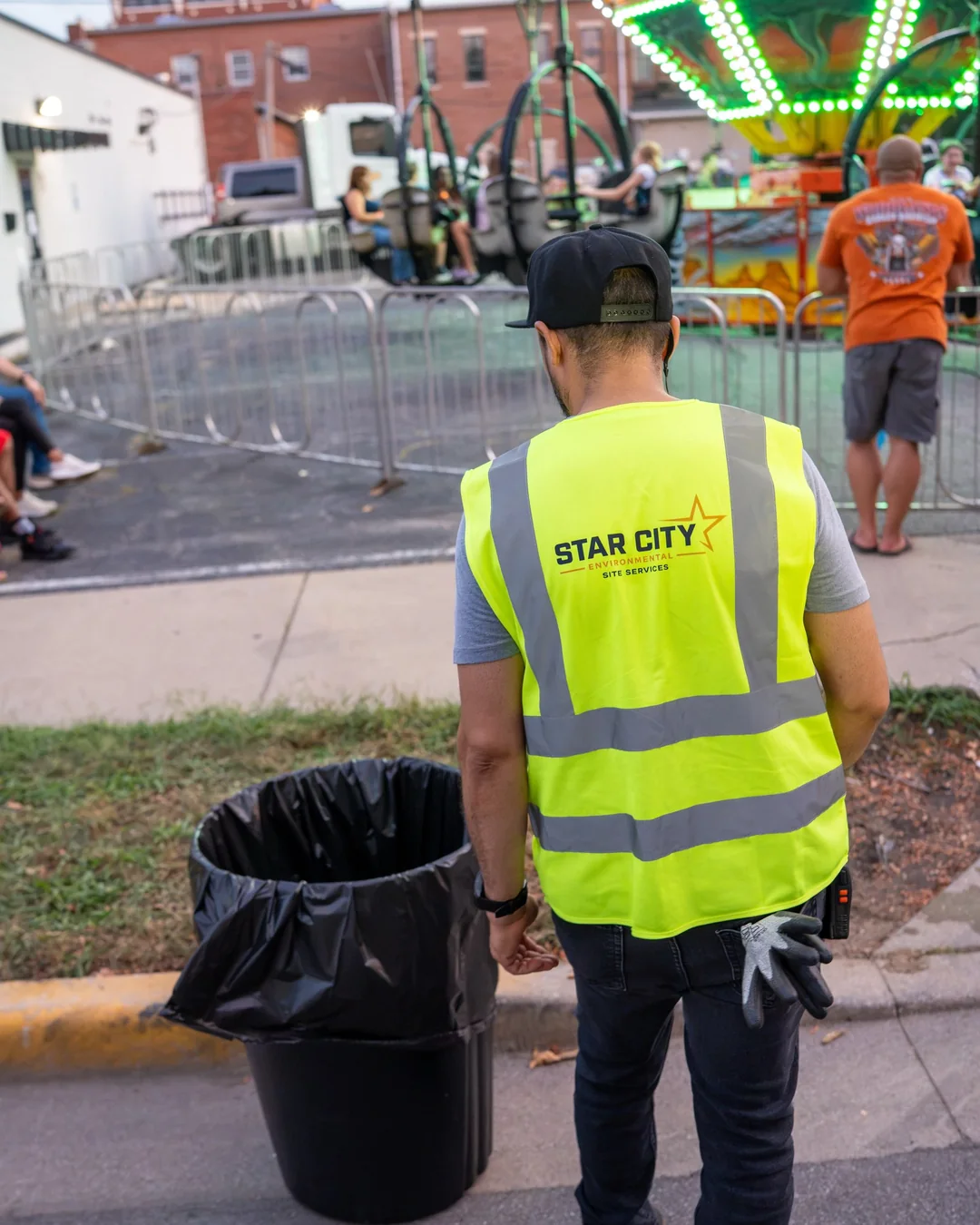 A crew member in a Star City Environmental vest standing beside a lined trash can at the edge of a midway ride