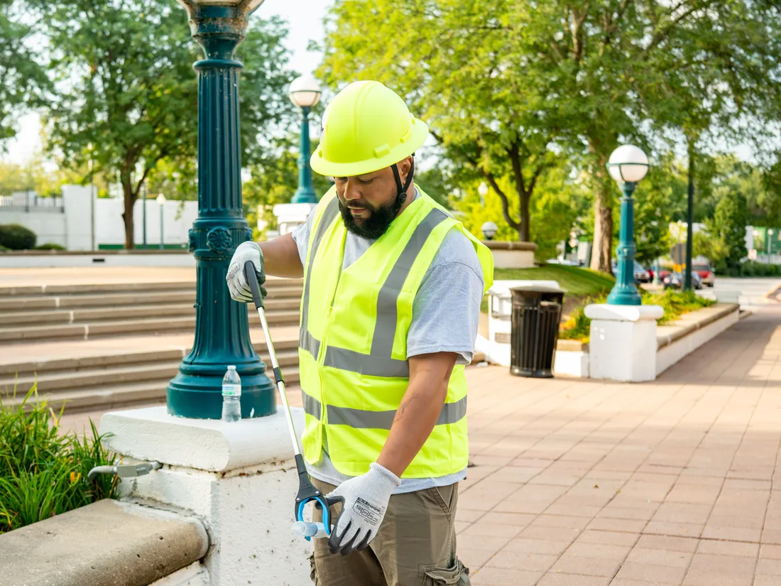 A Star City Environmental crew member in a hard hat and branded high-visibility vest servicing a receptacle on a downtown plaza.