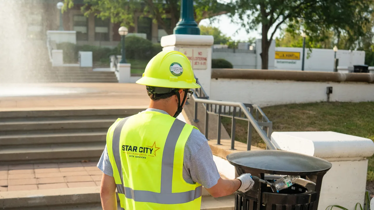 A crew member in a hard hat and high-visibility vest servicing a receptacle on a paved plaza.