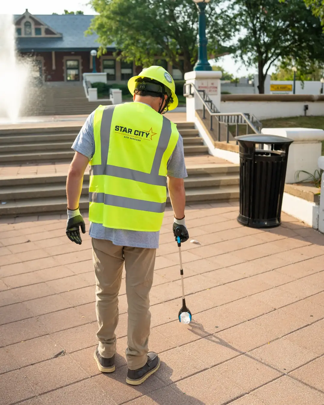 A crew member walking a plaza with a litter grabber beside a waste receptacle.
